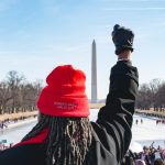 A woman stands on the steps of the Lincoln Memorial, facing the crowd and the Washington Monument, holding her fist up, and wearing a knitted cap with "Women's March Jan. 20, 2018" imprinted on it. People gathered around the reflecting pool for the "Women's March on Washington 2018", on Saturday, January 20, 2018. (Photo by Cheriss May) (Photo by Cheriss May/NurPhoto)