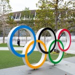 TOKYO, JAPAN, MARCH 27TH 2020 - Am man wearing protective mask stands by the Olympic logo at the new National Stadium built for the 2020 Olympics that have just be posponed to 2021 due to coronavirus outbreak