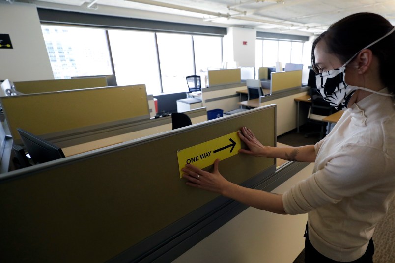 HOLD FOR STORY BY KELVIN CHAN — In this Thursday, May  7, 2020 photo, Interior Designer Stephanie Jones at the design firm Bergmeyer, applies a safe distancing reminder indicating one way foot traffic to a cubicle at the firms offices, in Boston. Out of concern for the coronavirus Bergmeyer is restructuring the way its workspace is used, including reinstalling dividers on 85 desks at its Boston office that had been removed over the years. (AP Photo/Steven Senne)